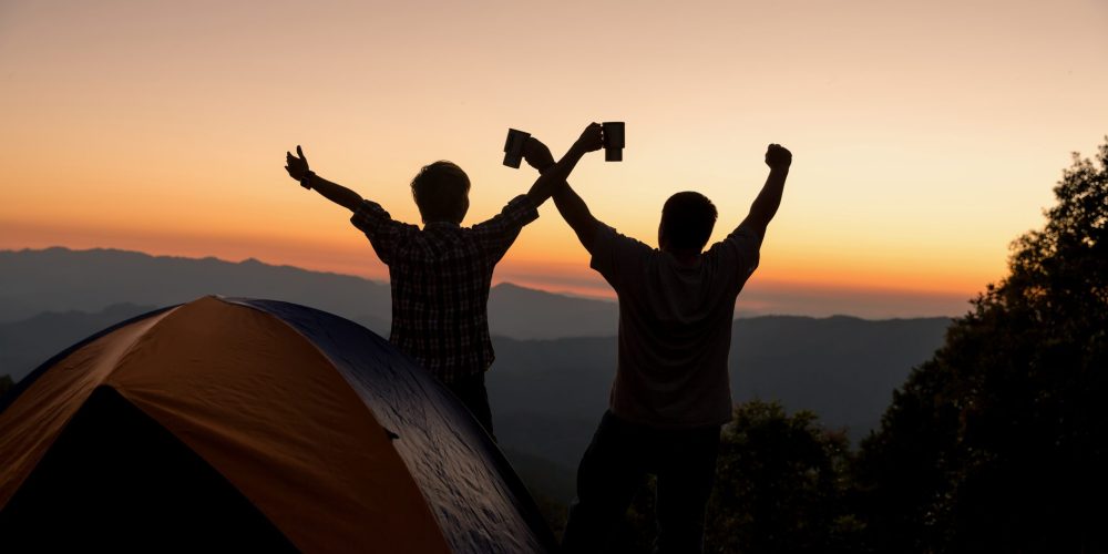 Two men tourists happy on the top of the mountain at near campfire under amazing sunset evening sky in a mountains area.